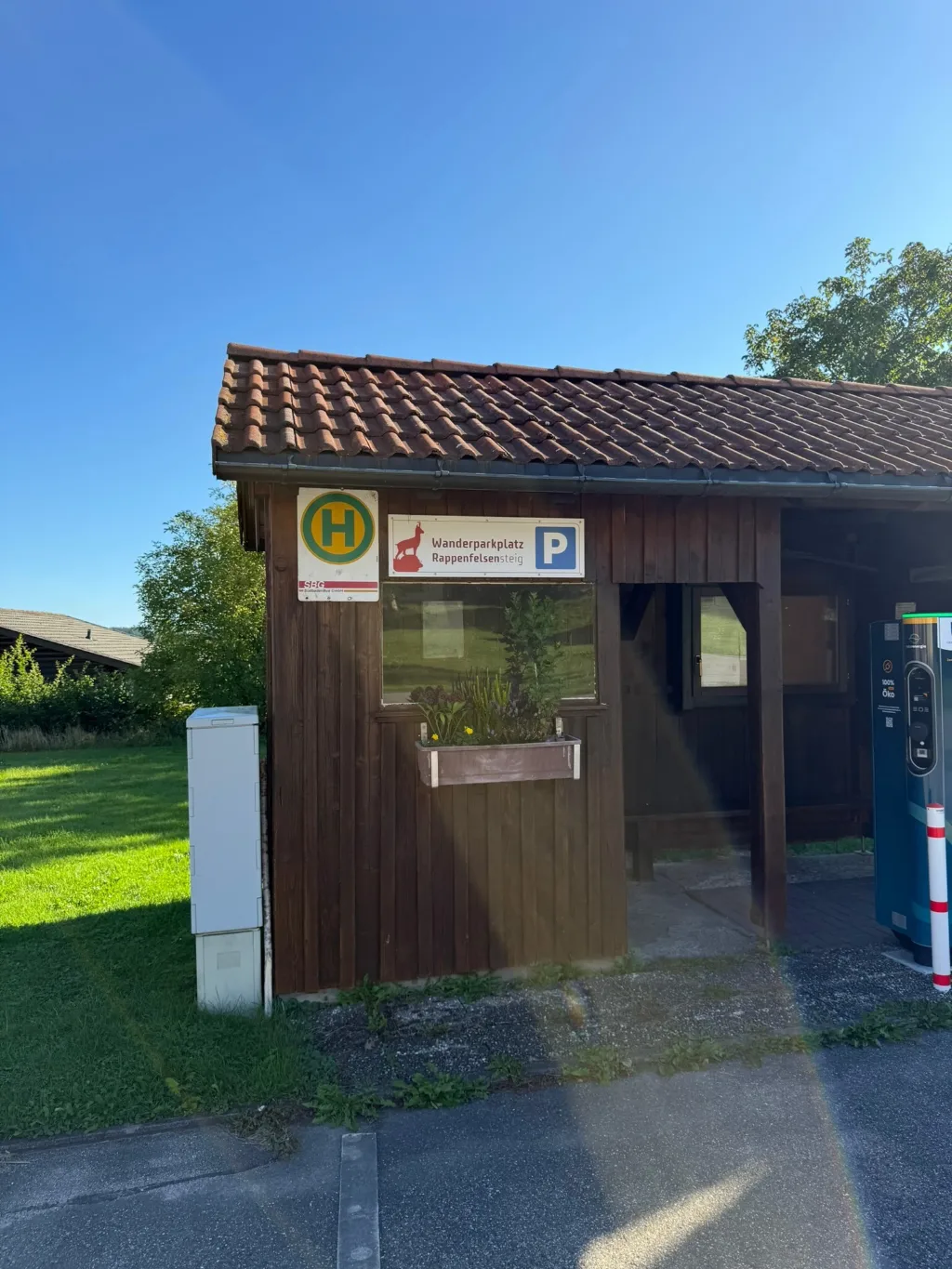 Hölzernes Bushaltestellenhäuschen mit Haltestellenzeichen und Schild „Wanderparkplatz Rappenfelsensteig“, daneben Ladesäule.