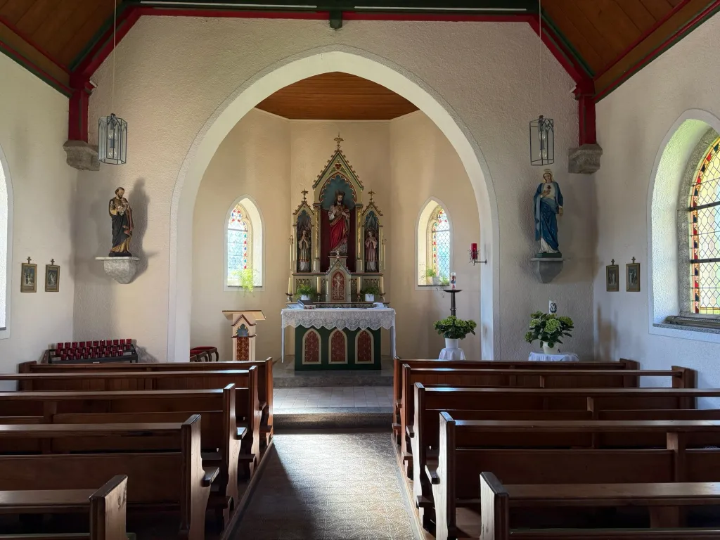 Blick durch den Mittelgang einer kleinen Kapelle mit Holzbänken zum bunten Altar mit Heiligenfiguren.