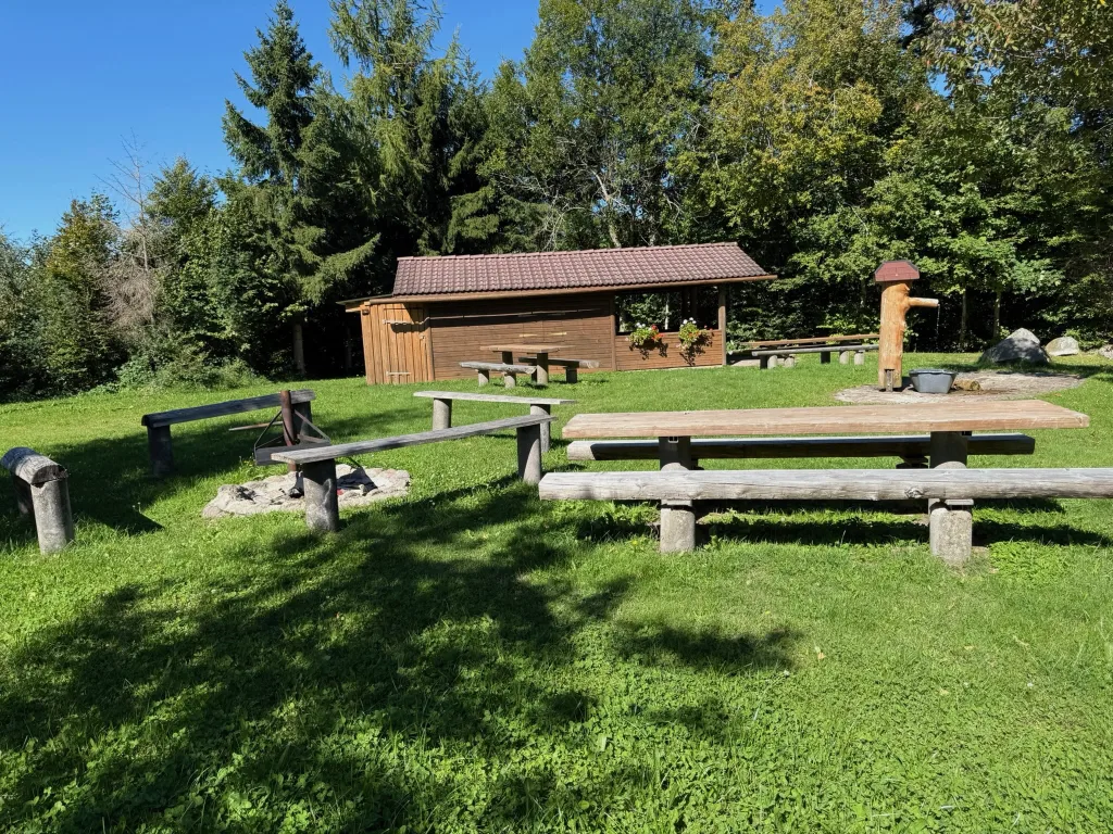Grillplatz auf einer Wiese mit Feuerstelle, Holztischen und Bänken vor einer Holzhütte am Waldrand.
