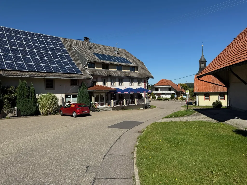 Dorfstraße mit Gasthaus, blauen Sonnenschirmen, rotem Auto und Solardach, rechts eine Kapelle mit Turm.