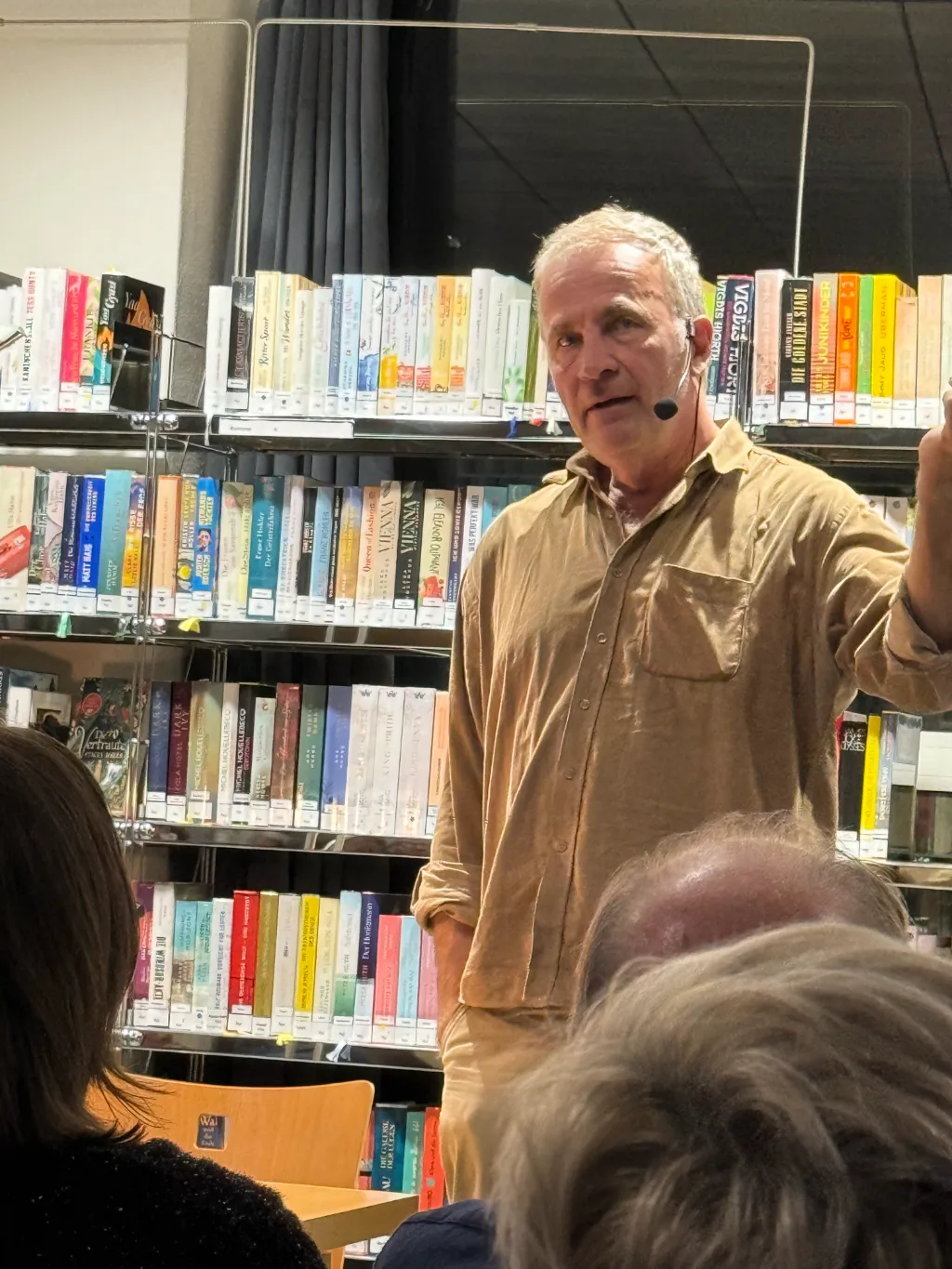 Alex Capus mit Headset-Mikrofon und erhobenem Arm beim Erzählen vor Bücherregalen in der Mediathek Wehr.