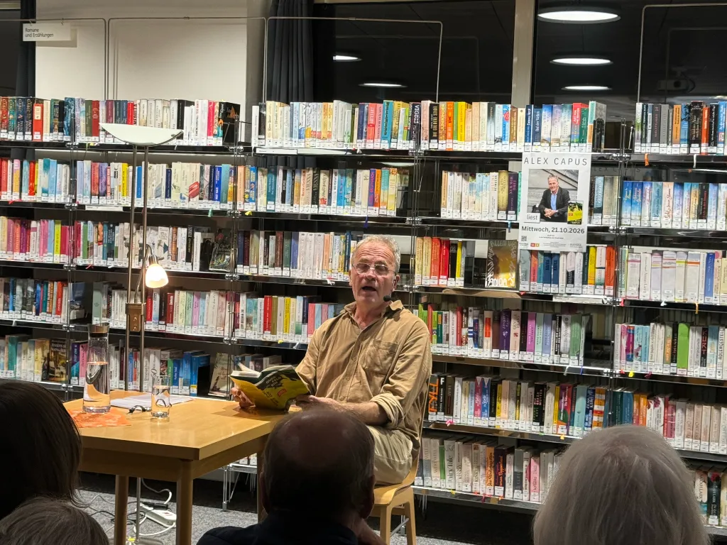 Alex Capus liest mit aufgeschlagenem Buch vor Bücherregalen in der Mediathek, daneben ein Tisch mit Lampe.