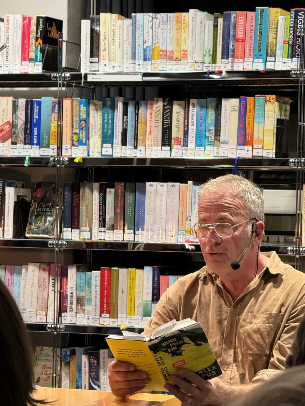 Alex Capus liest bei einer Lesung in der Mediathek vor einem Bücherregal aus seinem Buch „Querfeldein“.