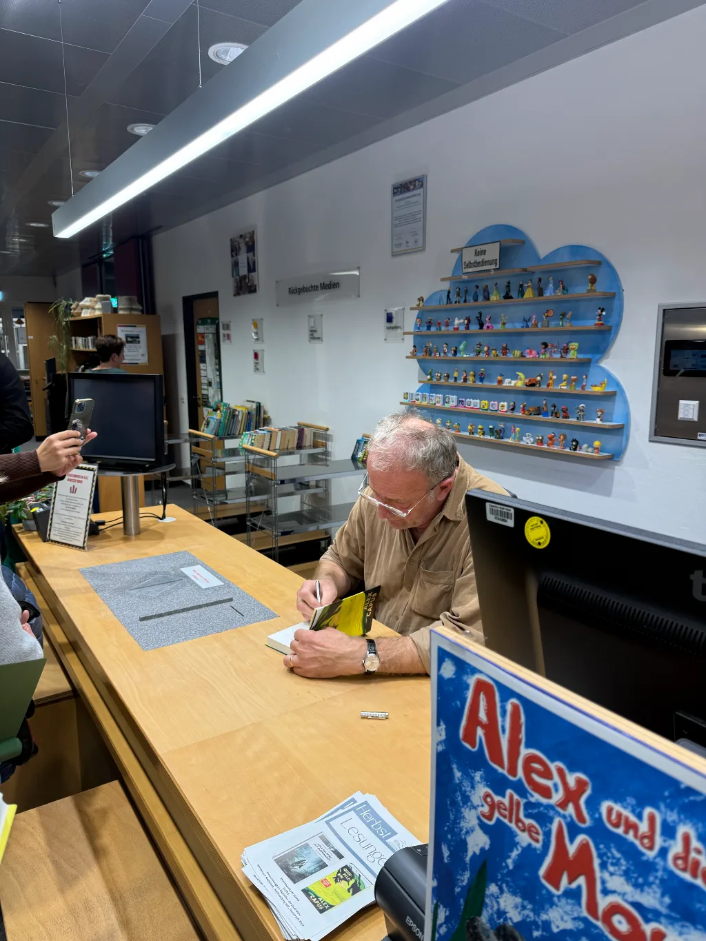 Alex Capus signiert nach der Lesung an der Theke der Mediathek Wehr ein Buch, davor ein Plakat.
