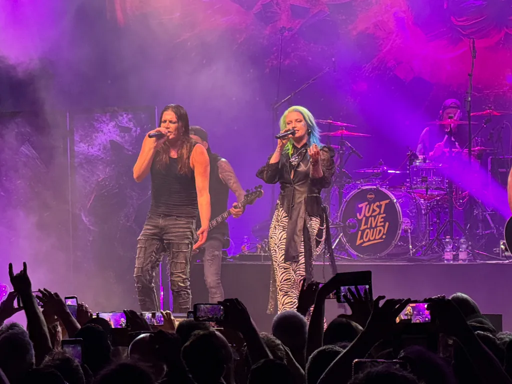 Mark Fox und Seraina Telli singen auf einer violett beleuchteten Bühne vor Publikum, Schlagzeug mit Aufschrift "Just Live Loud!".