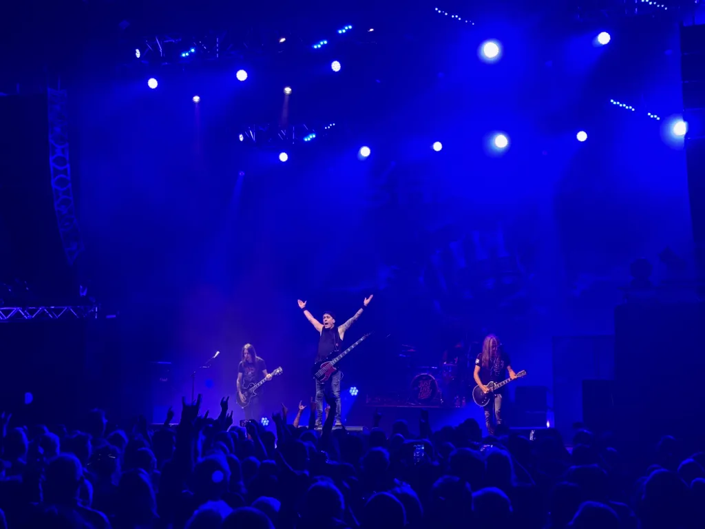 Drei Musiker auf blau ausgeleuchteter Bühne, der Bassist reißt die Arme hoch, davor jubelndes Publikum.