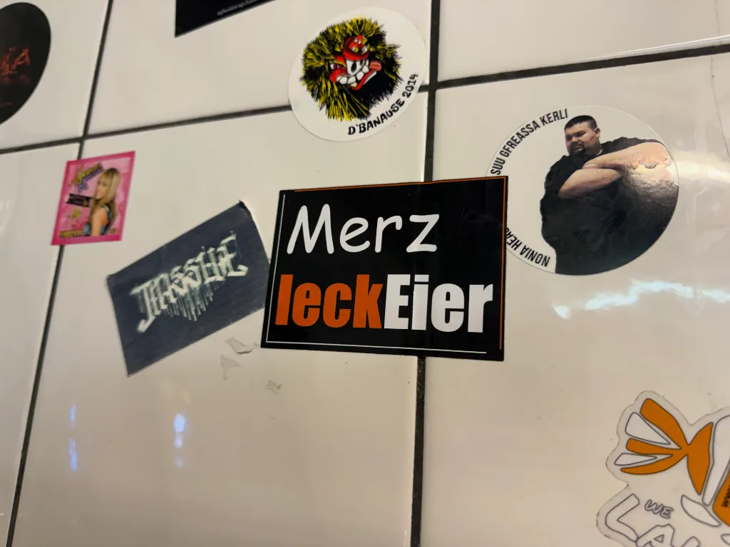 Mit Stickern beklebte weiße Kachelwand, zentral ein schwarzer Aufkleber mit der Aufschrift „Merz leck Eier“.