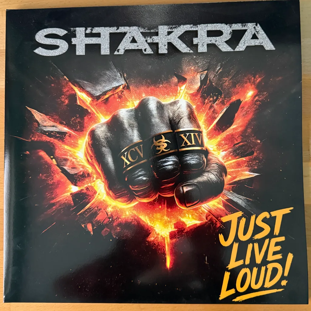 Albumcover von Shakra „Just Live Loud!“ mit geballter Faust vor Feuerexplosion, als Vinyl-Ausgabe auf Holztisch