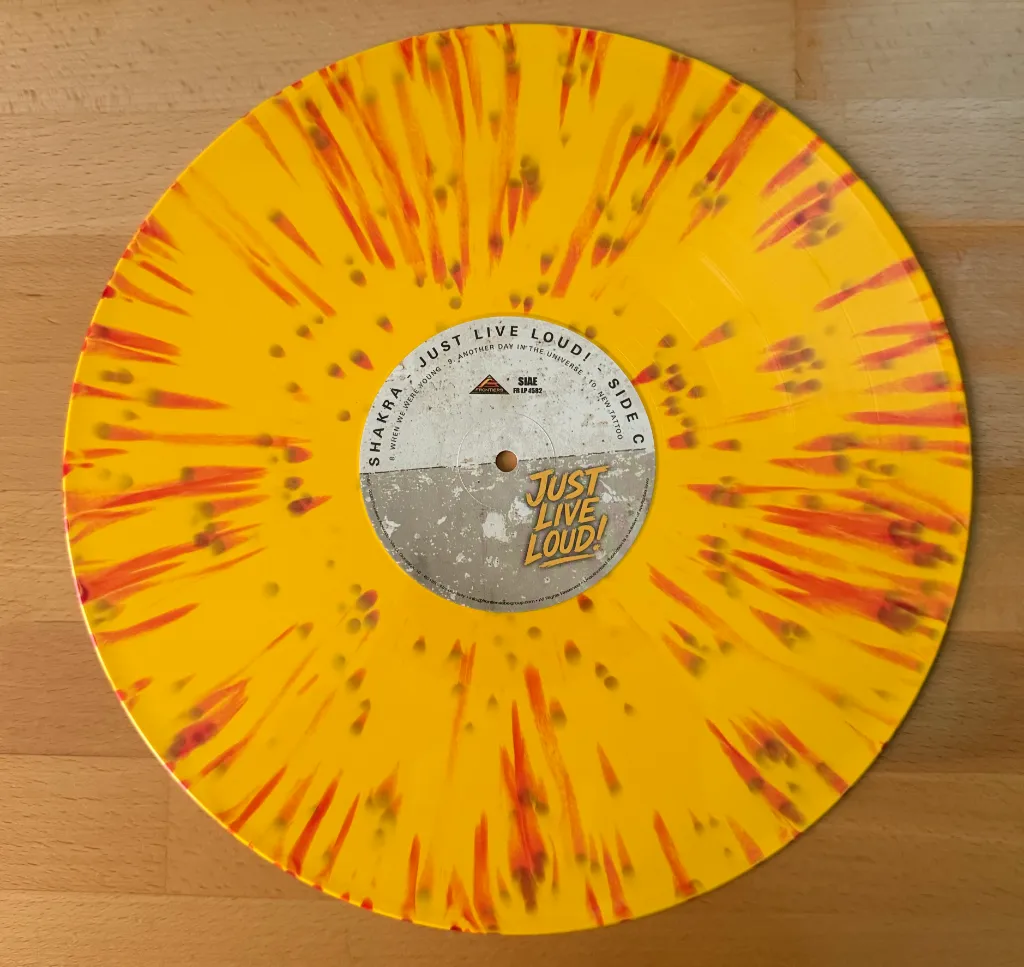 Gelbe Vinyl-Schallplatte mit roten Spritzern, Label „Shakra – Just Live Loud! Side C“, auf hellem Holztisch.