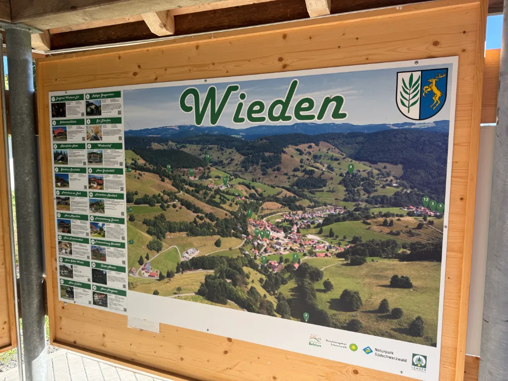 Informationstafel mit Luftbild des Schwarzwalddorfs Wieden, Ortswappen und Übersicht von Gasthöfen in einer Holzhütte.