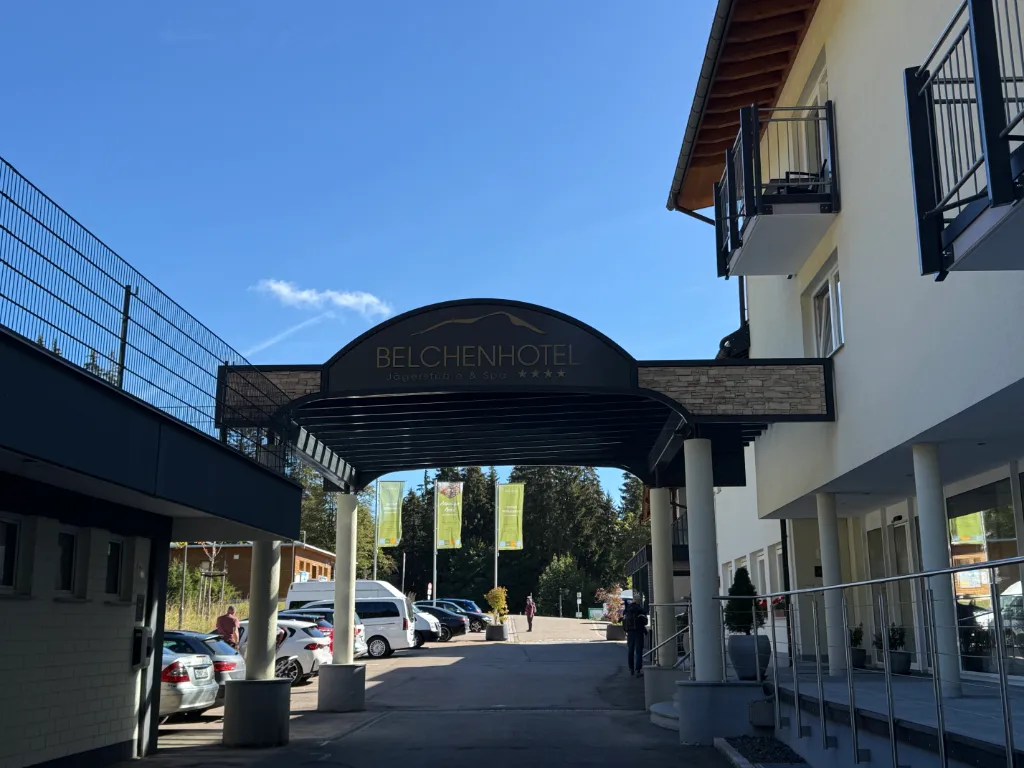 Überdachte Einfahrt des Belchenhotels mit geschwungenem Schriftzug, Autos auf dem Parkplatz unter blauem Himmel.