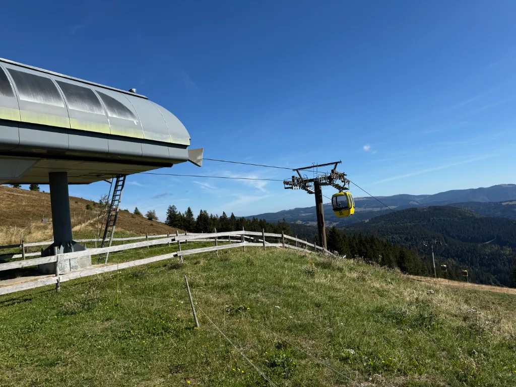 Bergstation der Belchen-Seilbahn mit gelber Gondel am Seil über einer Wiese vor blauem Himmel und Schwarzwaldpanorama