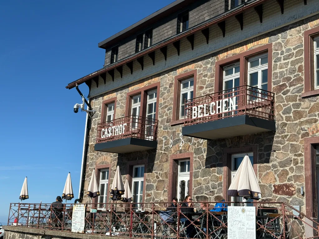 Steinfassade des Gasthof Belchen mit Balkonen und Schriftzug, davor Gäste an Tischen auf der sonnigen Terrasse.