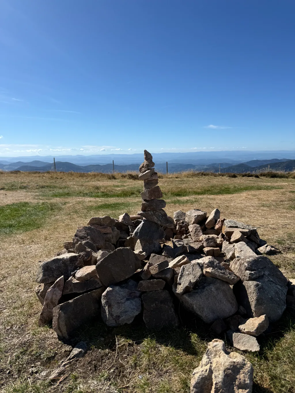 Steinmännchen auf einem Steinhaufen am Belchen-Gipfel, dahinter Wiese und weite Berglandschaft unter blauem Himmel.