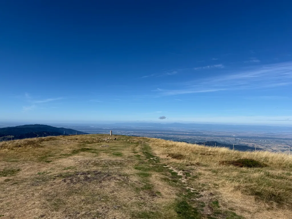 Weite Gipfelfläche des Belchen mit trockenem Gras, Trampelpfad und Fernsicht über die Rheinebene bei blauem Himmel