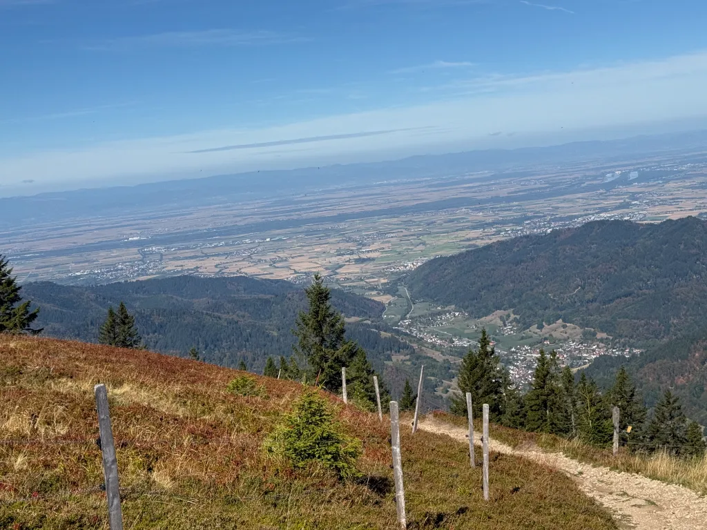 Weiter Blick vom Belchen über bewaldete Hänge und ein Dorf hinab in die Rheinebene, im Vordergrund ein Weidezaun.