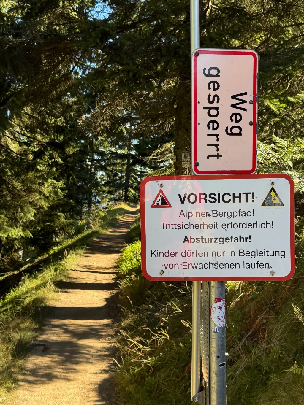 Schilder „Weg gesperrt“ und „Vorsicht! Alpiner Bergpfad“ an einem Mast neben einem schmalen Waldpfad.