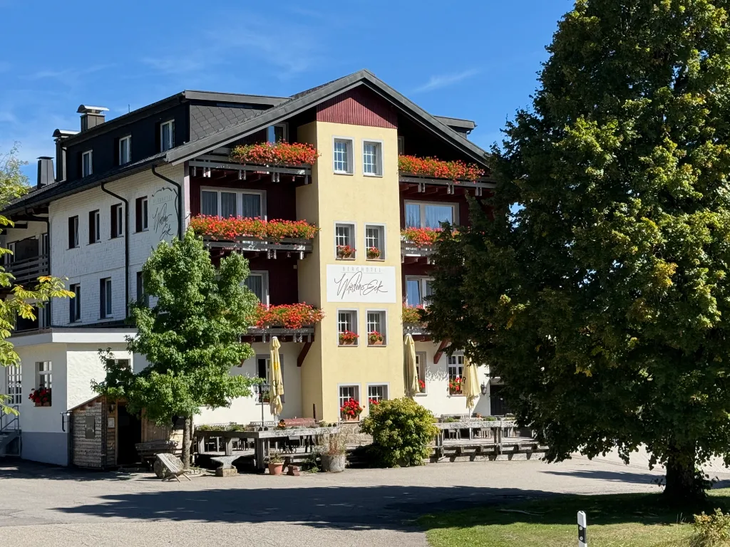 Mehrstöckiges Gasthaus mit rot-gelber Fassade, Blumenkästen und Terrasse, daneben ein großer Laubbaum