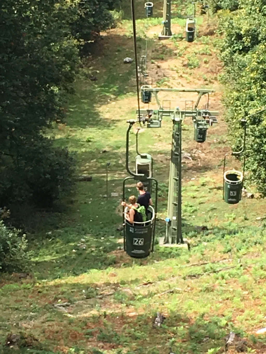 Zwei Personen fahren in einer offenen grünen Seilbahn-Gondel mit der Nummer 26 einen bewaldeten Hang hinauf.