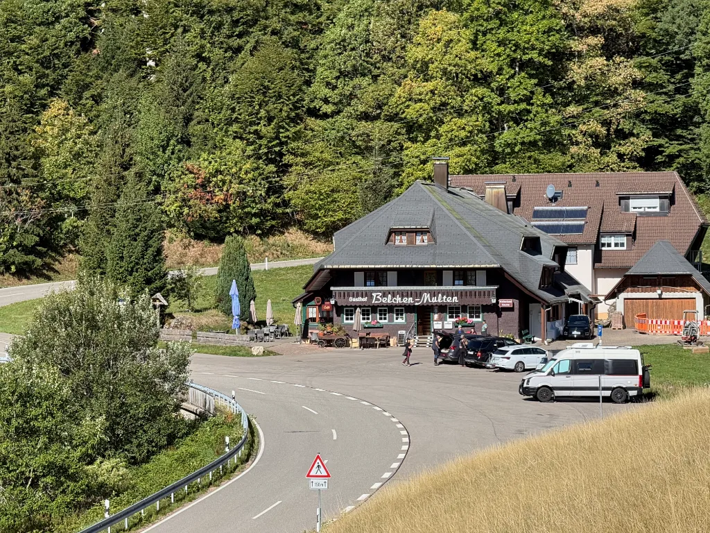 Der Gasthof Belchen-Multen, ein Schwarzwaldhaus mit großem Walmdach, liegt an einer Straßenkurve am bewaldeten Hang.