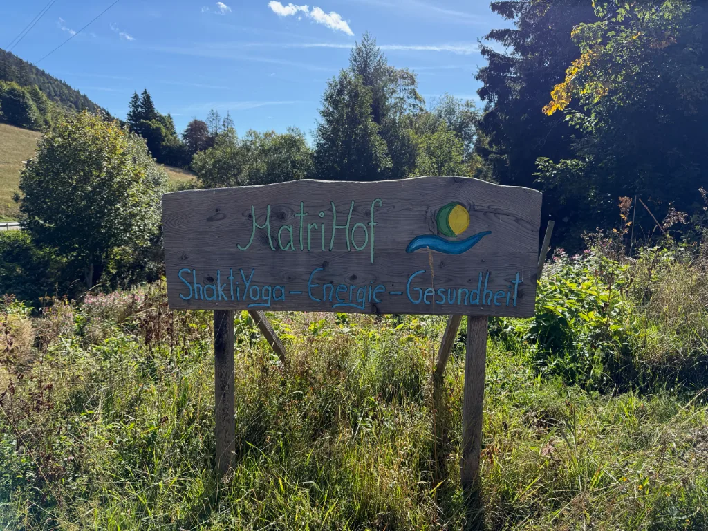 Handgeschnitztes Holzschild am Wegrand mit der Aufschrift „MatriHof – ShaktiYoga – Energie – Gesundheit“ in hoher Wiese.